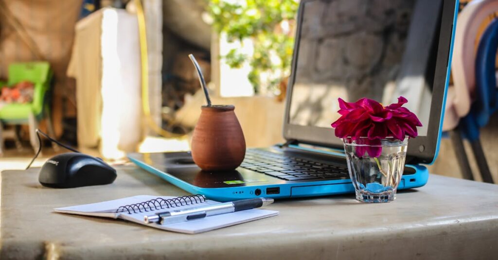 A serene outdoor workspace featuring a laptop, notebook, mug, and flowers on a desk.